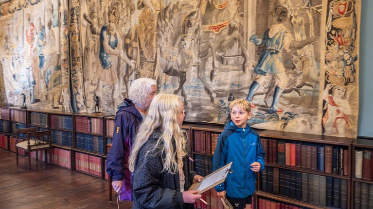 Two blonde haired children and their grandmother walking through the house at Sizergh. They are talking animatedly and walking past a large tapestry.